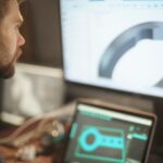A man wearing a black collard shirt sits at a desk, working on a design rendering on a laptop screen and desktop screen on the desk in front of him.